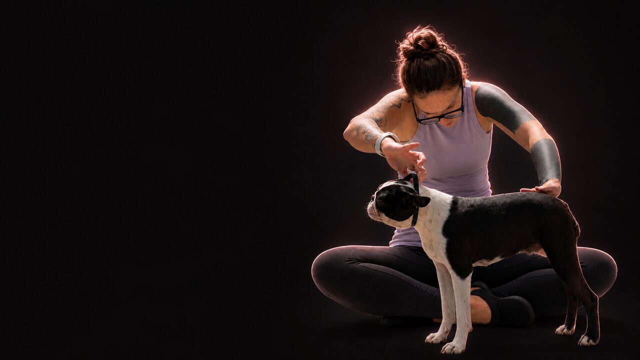 Educatrice cinofila seduta a gambe incrociate mentre lavora con un cane, avvolta da una luce calda su fondo nero